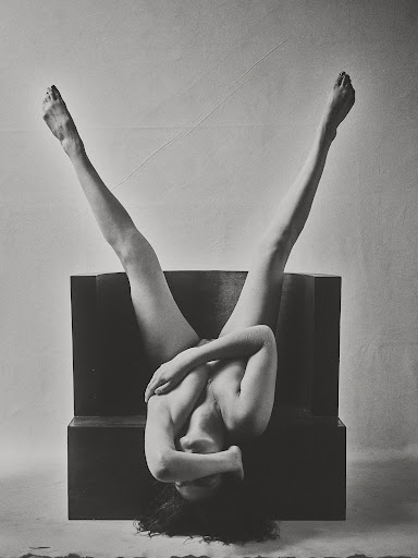 Monochrome photograph of an adult female upside down on a dark block, legs extended, face obscured, arms crossing her chest in an indoor setting.
