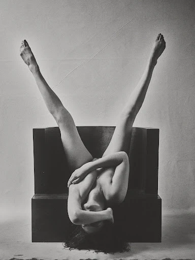 Monochrome photograph of an adult female upside down on a dark block, legs extended, face obscured, arms crossing her chest in an indoor setting.