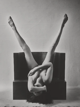 Monochrome photograph of an adult female upside down on a dark block, legs extended, face obscured, arms crossing her chest in an indoor setting.