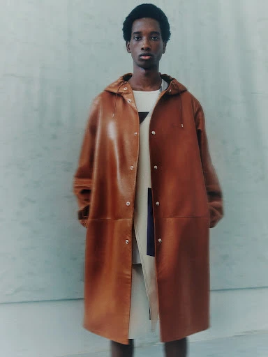 Adult male model poses in a hooded, knee-length brown leather coat over a white top against a neutral pale-blue plaster background.