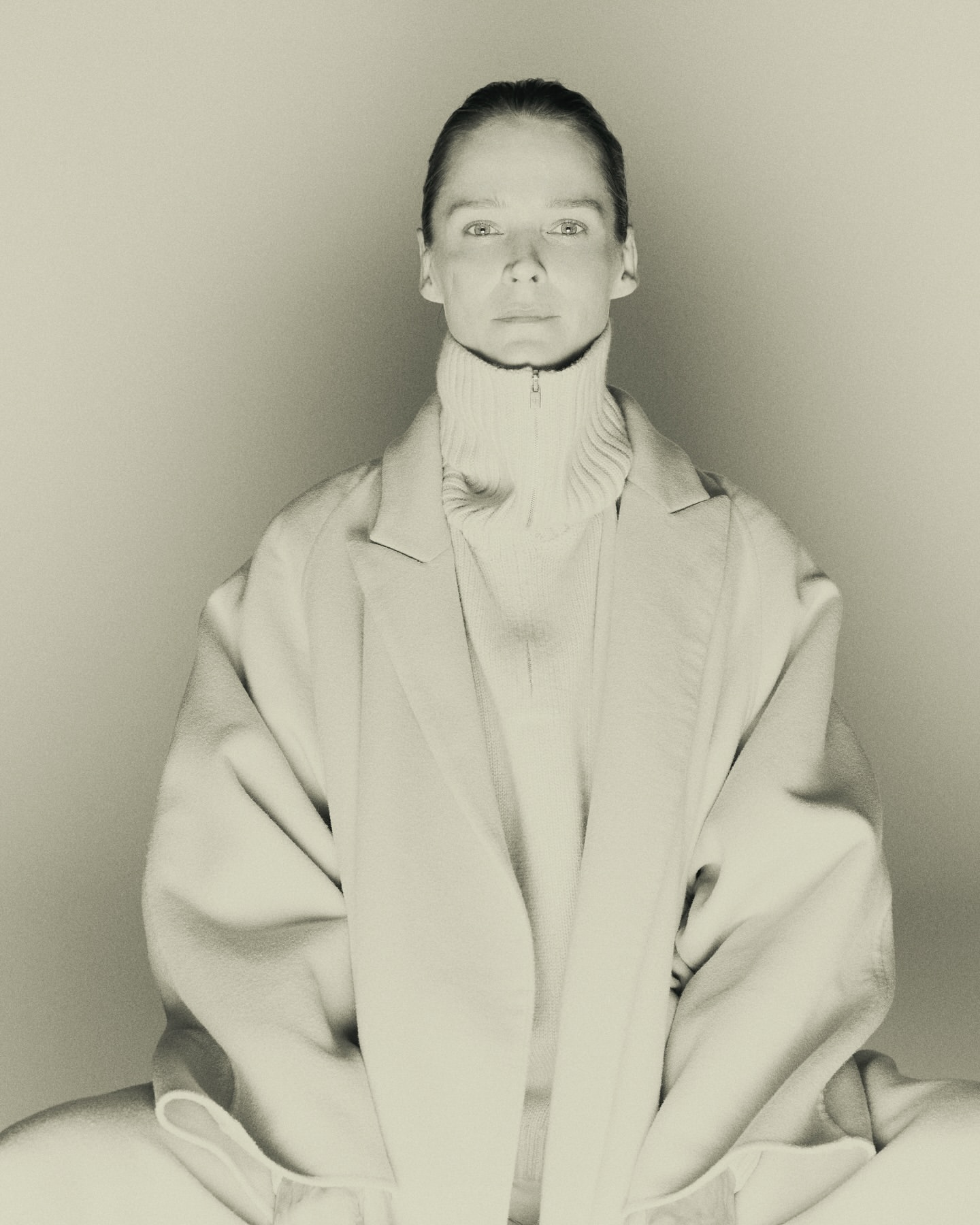 Monochrome portrait of a model wearing a high-collared knit sweater and oversized coat against a neutral background