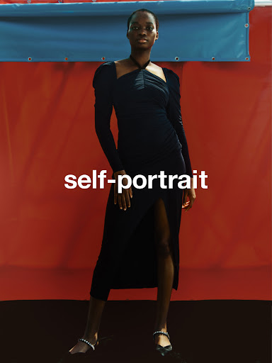 A Black female fashion model poses indoors against an orange wall wearing a long, black dress and heeled sandals with embellishments.