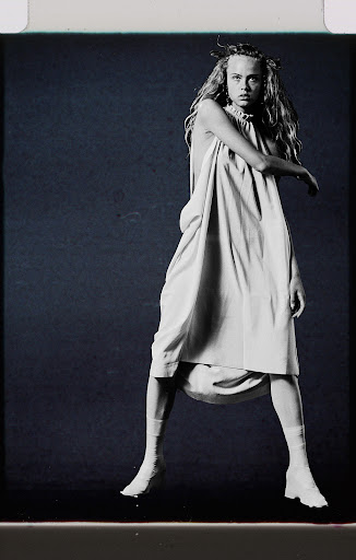 A young adult woman with long blonde hair poses in a studio wearing a pale dress, white socks, and shoes against a dark background.