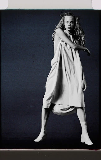 A young adult woman with long blonde hair poses in a studio wearing a pale dress, white socks, and shoes against a dark background.