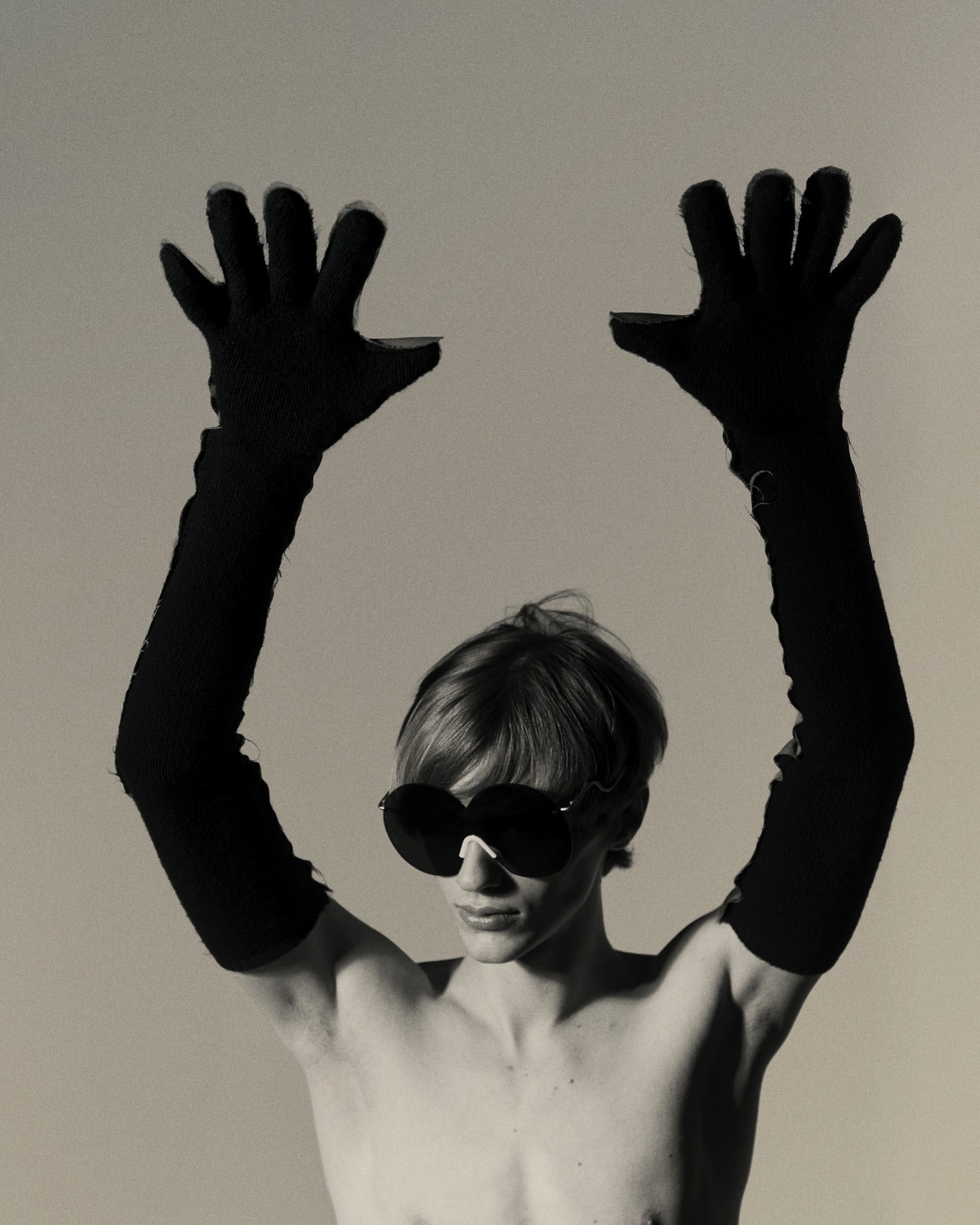 Black and white portrait of a bare-chested person wearing large sunglasses and long gloves, holding their hands up in a photo studio.