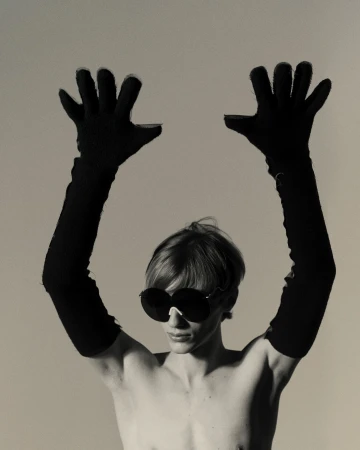 Black and white portrait of a bare-chested person wearing large sunglasses and long gloves, holding their hands up in a photo studio.