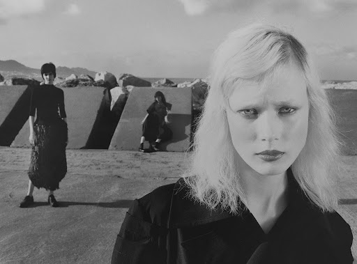Black and white fashion shot featuring a model in the foreground and two models in the background by concrete structures near a coastline.