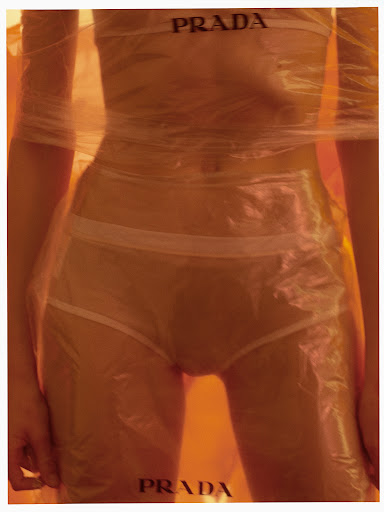Fashion shot of a person wrapped in clear plastic, wearing white underwear, with the brand name PRADA appearing at the top and bottom.