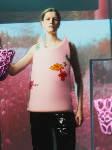 Adult female model wears pink floral top and latex pants in a studio setting with projected background imagery and prop.