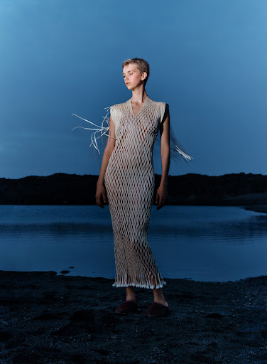 Young woman in a mesh dress poses in a dimly lit outdoor setting with a lake and dark hills in the background under a dusk sky.