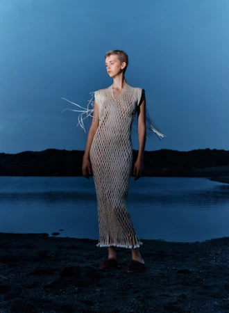 Young woman in a mesh dress poses in a dimly lit outdoor setting with a lake and dark hills in the background under a dusk sky.