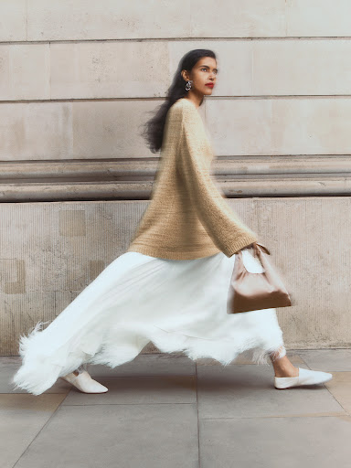 Woman walking past a stone building wearing a tan sweater, white pleated skirt, and holding a small handbag.