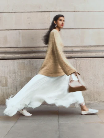 Woman walking past a stone building wearing a tan sweater, white pleated skirt, and holding a small handbag.
