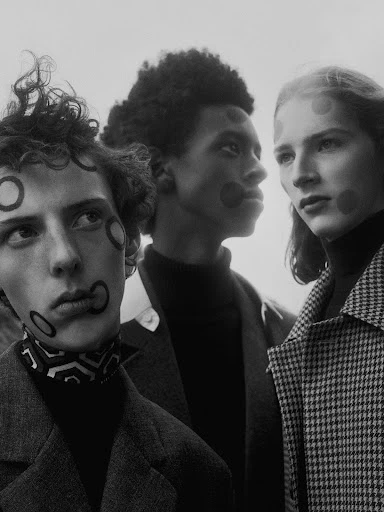 Three young adult fashion models in a studio shot. They wear dark turtlenecks, coats and patterned makeup. Black and white.