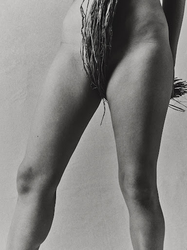 Monochrome photo of the lower half of a nude woman standing with her legs apart and raffia draped around her waist and thighs.
