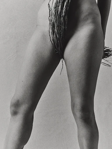 Monochrome photo of the lower half of a nude woman standing with her legs apart and raffia draped around her waist and thighs.