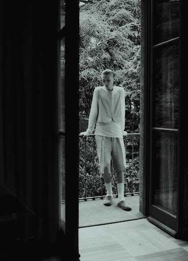 Model standing on a balcony leaning against a railing viewed from inside a room through open glass doors by Nicolas Kern