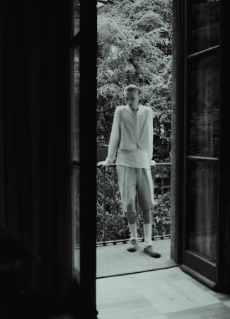 Model standing on a balcony leaning against a railing viewed from inside a room through open glass doors by Nicolas Kern