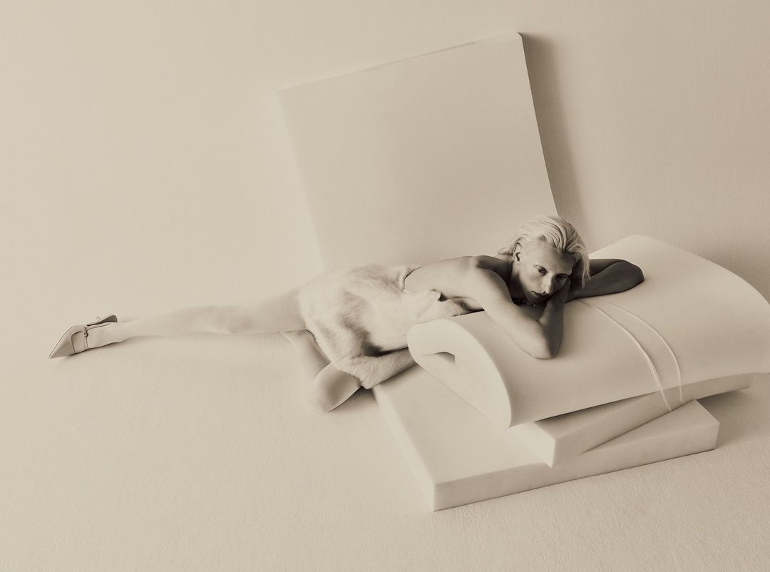Monochromatic image of a reclining adult woman with fair skin and blonde hair, posing in high heels on white geometric foam shapes.