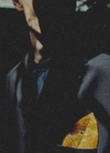 Close-up of an adult wearing a dark suit, dress shirt, and pocket square. The man's face is obscured by shadow.
