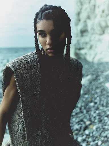 Young woman with braided hair poses in textured knit dress on stony beach with chalk cliffs and overcast sky.