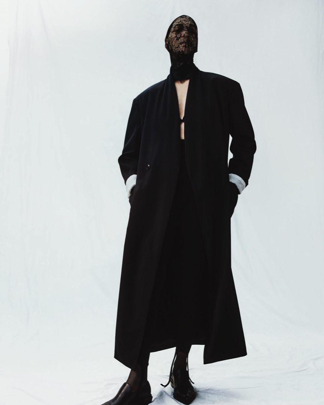 Fashion-forward individual in a sleek black coat and lace mask against a clean white backdrop, highlighting a modern, edgy aesthetic.