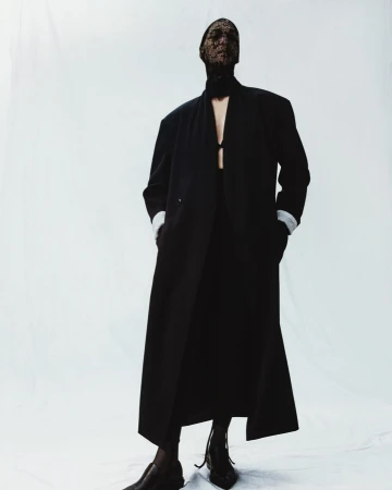 Fashion-forward individual in a sleek black coat and lace mask against a clean white backdrop, highlighting a modern, edgy aesthetic.