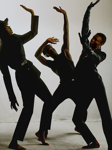 Three black models in dark attire posing dynamically with raised arms against a white seamless studio backdrop.