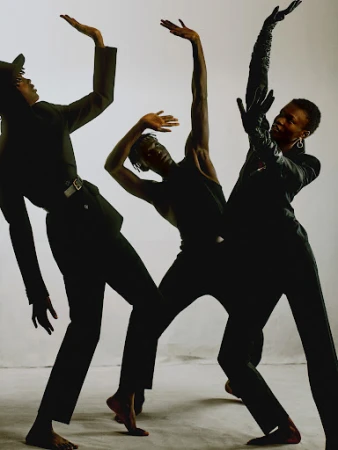 Three black models in dark attire posing dynamically with raised arms against a white seamless studio backdrop.