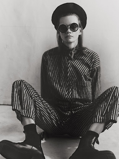 Model wearing round sunglasses, beret, and striped outfit seated against a plain backdrop