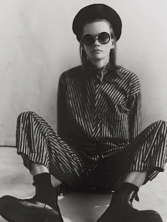 Model wearing round sunglasses, beret, and striped outfit seated against a plain backdrop