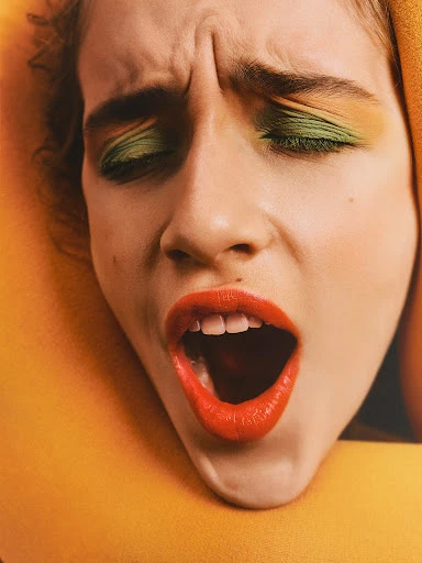 Close-up studio shot of an adult woman with green eye shadow and red lipstick, looking surprised, framed by a yellow object.