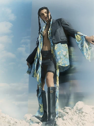Fashion shot of a person in a blazer, shorts, floral top, and high boots posing on a rock pile under a partly cloudy sky.