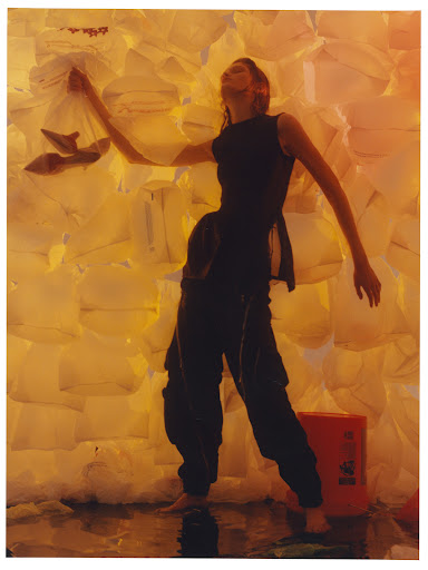 A woman in dark clothing and bare feet stands in water, posing in front of a wall made of illuminated white plastic bags.
