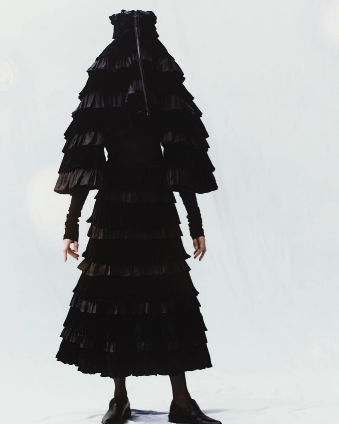 Fashion shoot featuring an unrecognizable person completely covered in a dramatic, multi-tiered black ruffle dress against a stark white backdrop.