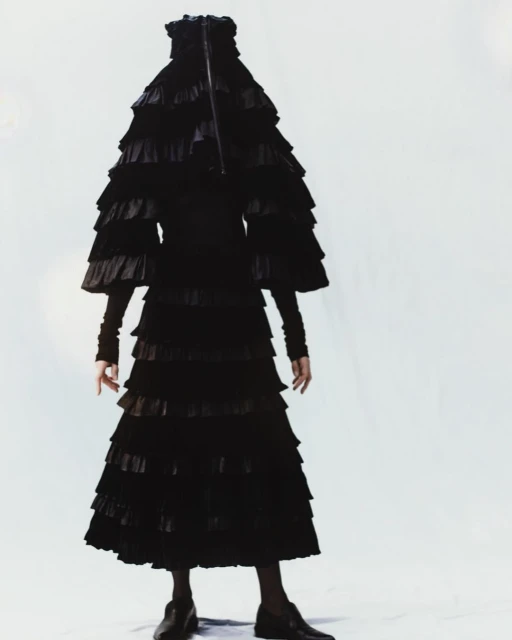 Fashion shoot featuring an unrecognizable person completely covered in a dramatic, multi-tiered black ruffle dress against a stark white backdrop.