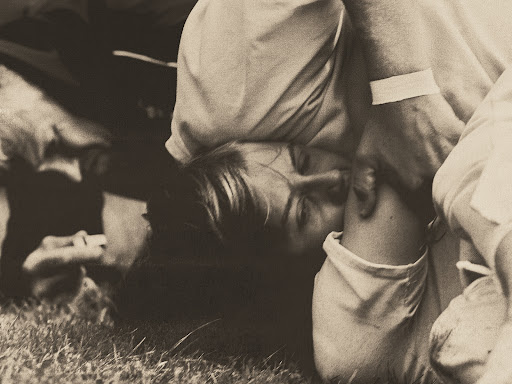 Monochromatic image of two adults, one lying on the ground with a bandage on their arm. A close-up shot captures the solemn scene.