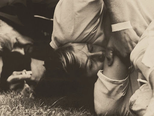 Monochromatic image of two adults, one lying on the ground with a bandage on their arm. A close-up shot captures the solemn scene.