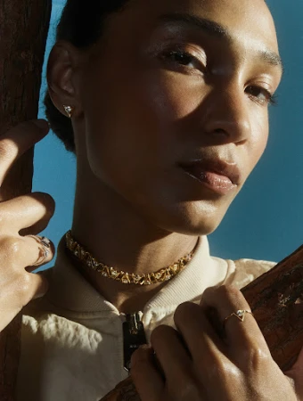 Close-up portrait of a woman wearing gold jewellery including a choker necklace, earrings, and rings against a blue sky.