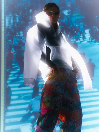 A young woman poses in a sheer hooded top and colorful pants, standing against a blurred blue backdrop of a crowded city scene.