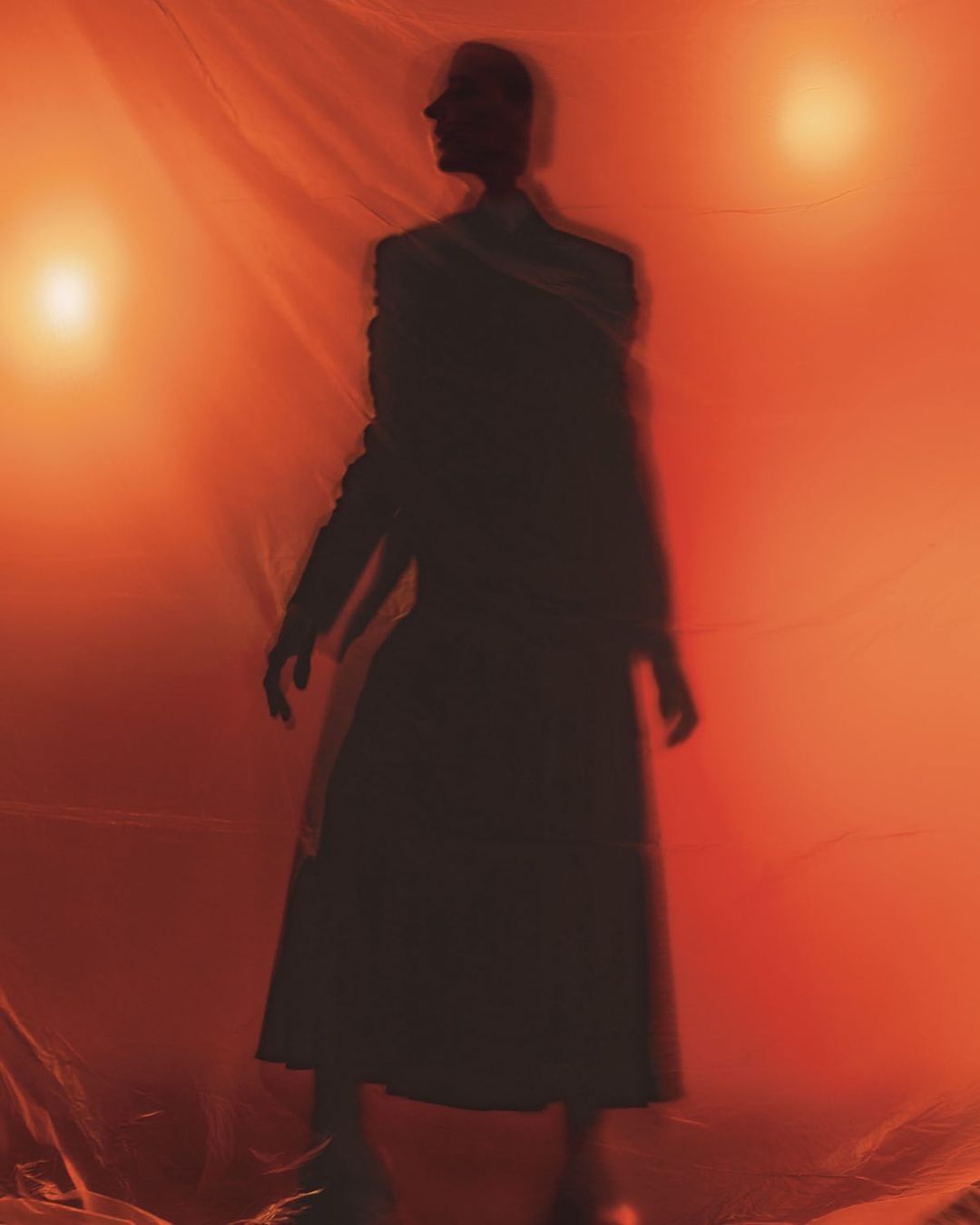 Silhouette of a person in a flowing garment stands against an orange backdrop with soft lighting, creating an ethereal, obscured figure.