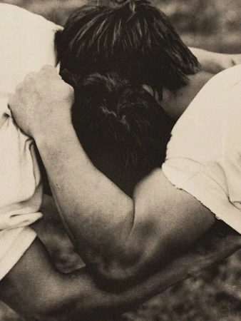 Close-up portrait of two people embracing closely with dark hair and white t-shirts in grainy black and white photography.