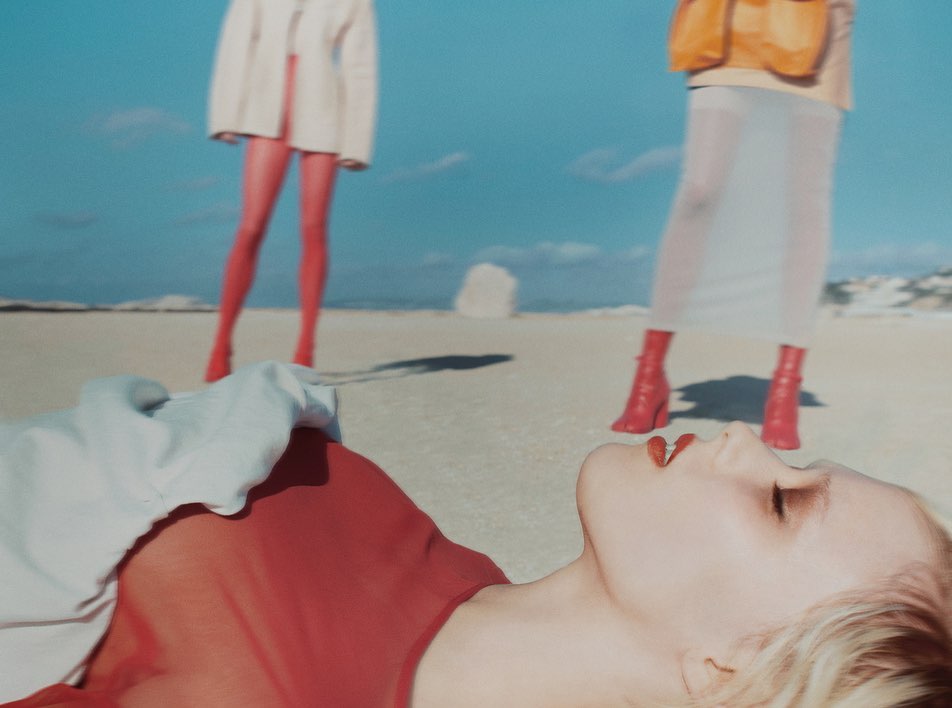 Model lying on sandy beach in red top with two standing figures in background under clear blue sky.