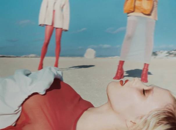 Model lying on sandy beach in red top with two standing figures in background under clear blue sky.