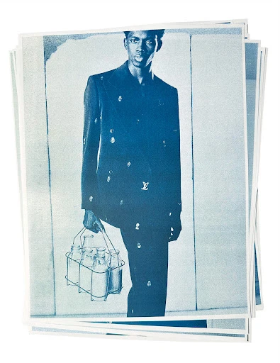 Cyanotype photo of a Black male model in a suit holding a metal carrier with glass milk bottles, against an abstract white background.