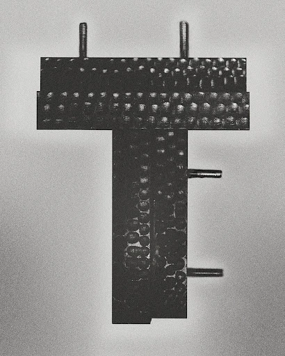 Close-up of a black metal T-shaped industrial component with bolts against a gradient gray background in a studio setting.