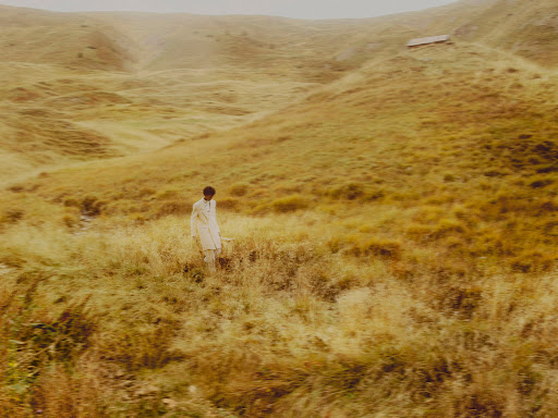 Person walking in a grassy field with rolling hills in the background, wearing a light-colored outfit in a natural light setting.