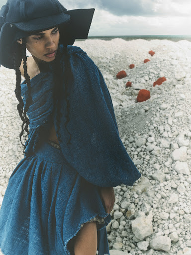 Woman in draped blue fabric outfit and hat poses in a rocky, outdoor, grey-toned environment under overcast sky in a fashion shot.