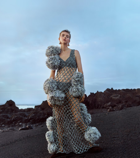 A young woman stands on a black sand beach wearing an avant-garde net dress adorned with grey pom-poms under a dusky sky at dusk.