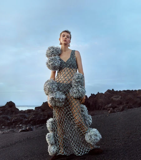 A young woman stands on a black sand beach wearing an avant-garde net dress adorned with grey pom-poms under a dusky sky at dusk.